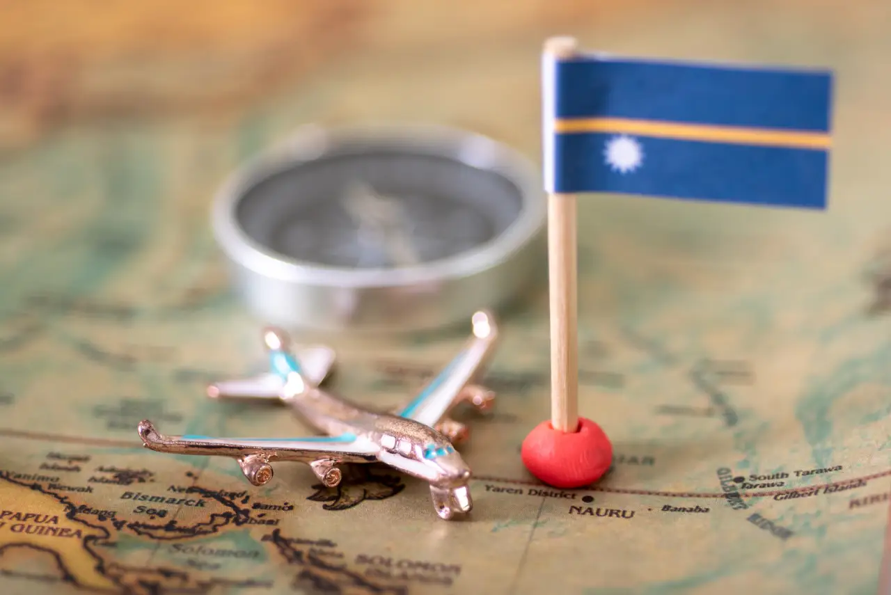 Would You Pay $105K For A Passport? This Island Hopes So 6 A small Nauru flag on a wooden stick, placed on a vintage map, with a miniature airplane and a compass in the background, symbolizing travel and citizenship.