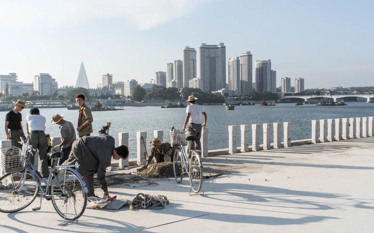North Korea Reopens Rason For Tourism After 5-Year Covid-19 Closure 2 Workers and cyclists by the Taedong River in Pyongyang, North Korea, with the city’s skyline and the Ryugyong Hotel visible in the background.