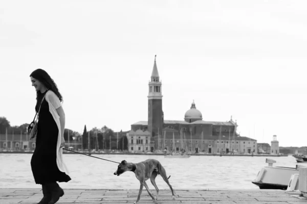 A woman in a flowing dress walks her dog along the waterfront in Venice, Italy, with the iconic San Giorgio Maggiore church in the background.