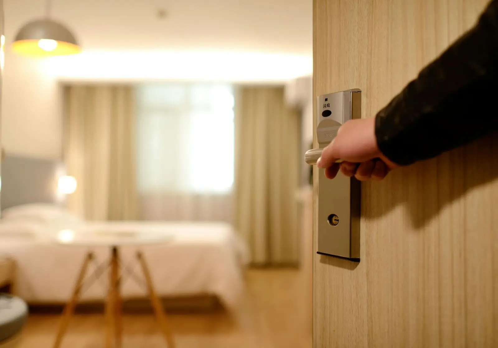 A hand opening a hotel room door with a cozy, well-lit bedroom in the background.