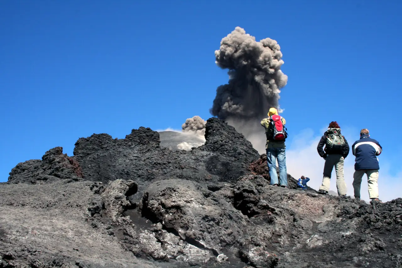 Tourists standing on rocky terrain near Mount Etna as it erupts, with a large plume of volcanic ash rising into the sky.