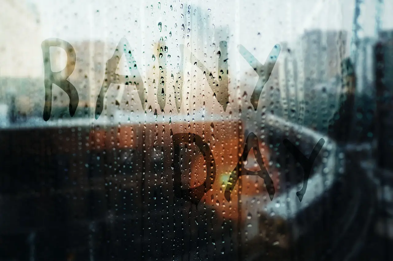 Jetblue Vacations Introduces First-Of-Its-Kind Rain Coverage 2 The words “RAINY DAY” written on a foggy, rain-covered window with a blurred cityscape in the background.