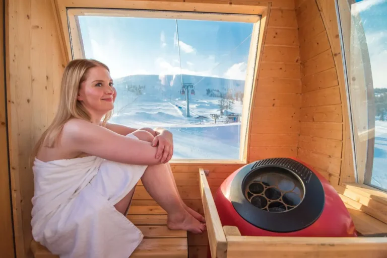 Other News 7 A woman enjoys the warmth of a sauna inside a wooden gondola lift, with a snowy ski resort visible through the large windows.