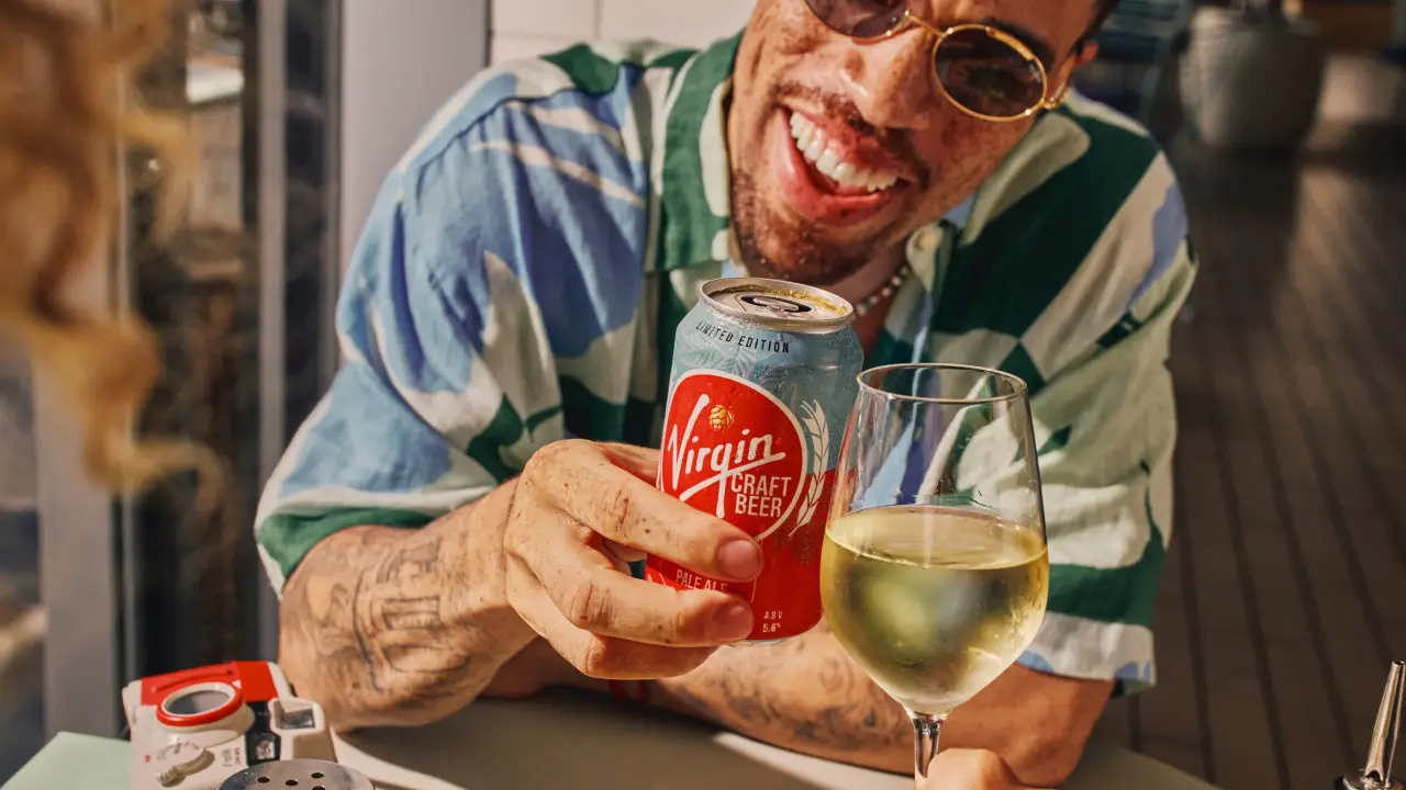 A man wearing sunglasses and a striped shirt smiles while holding a can of Virgin Craft Beer and a glass of white wine on a Virgin Voyages cruise.