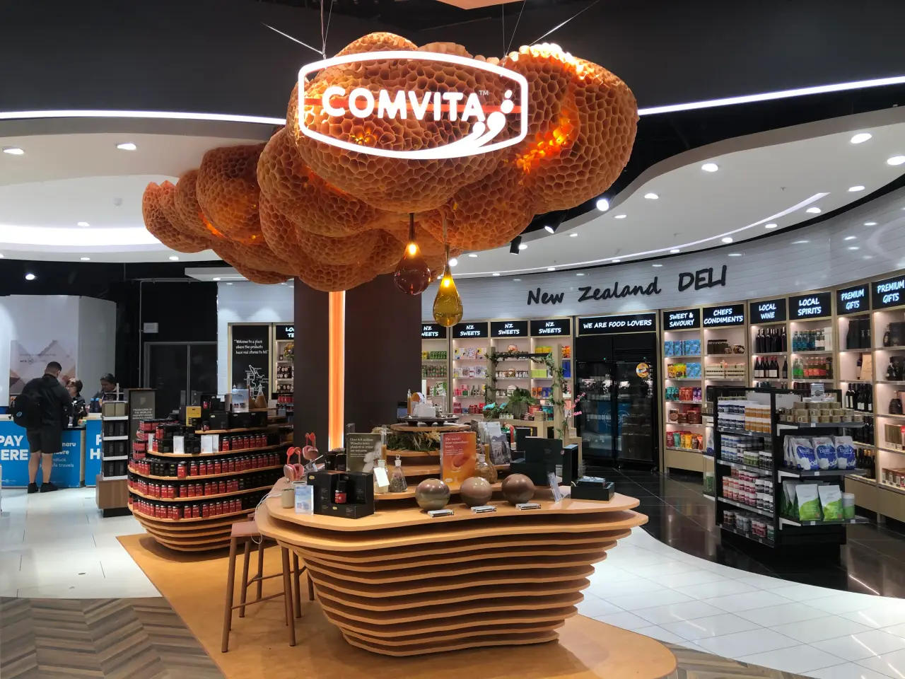 A Comvita honey-themed retail display at Auckland Airport, featuring a honeycomb-inspired ceiling design and a selection of honey products. The New Zealand Deli section is visible in the background, offering local sweets, wine, and premium gifts.