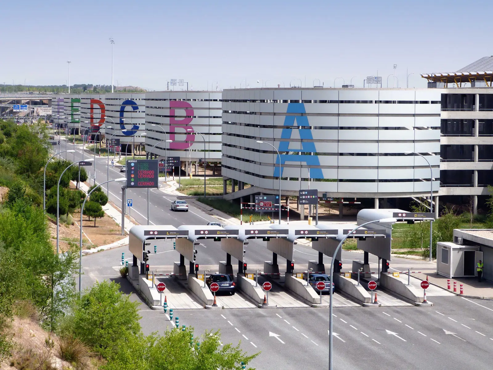 Madrid Airport parking entrance with toll booths and multi-level parking structures labeled with large colorful letters.