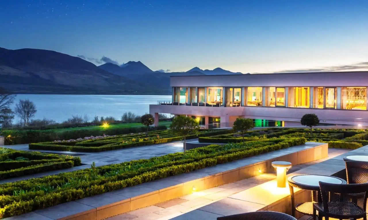 A luxury resort in Ireland with a beautifully landscaped terrace overlooking a serene lake and mountains at sunset, offering a tranquil and elegant setting.