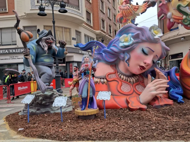 Spain Travel And Tourism News 19 Colorful Fallas sculptures in Valencia, Spain, featuring a sleeping woman with blue and purple hair, a wizard-like figure with a staff, and a warrior-like character. Spectators with umbrellas stand behind red barricades on a rainy day.