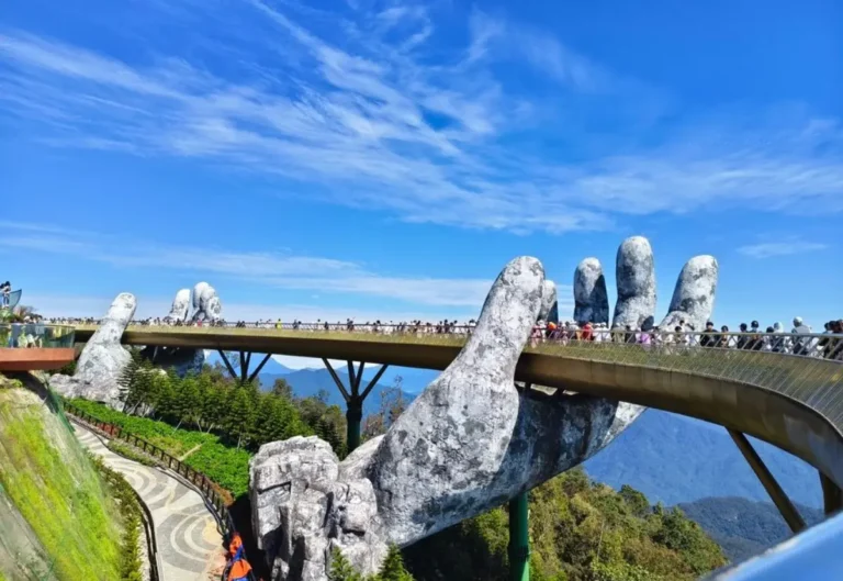 Golden Hands Bridge in Ba Na Hills, Vietnam, with tourists walking across, supported by giant stone-like hands against a backdrop of lush mountains and a clear blue sky.