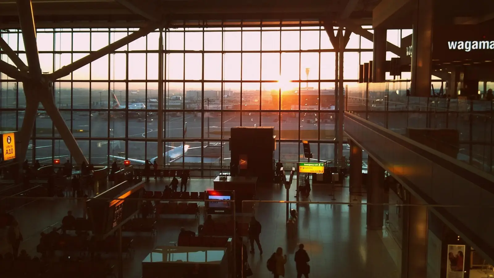 Heathrow Airport terminal with large glass windows overlooking the runway at sunset, with passengers waiting for flights.