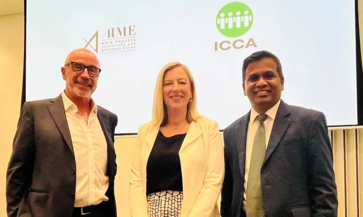 Three business professionals stand together at the ICCA and AIME partnership signing event, with the ICCA and AIME logos displayed on a screen behind them.