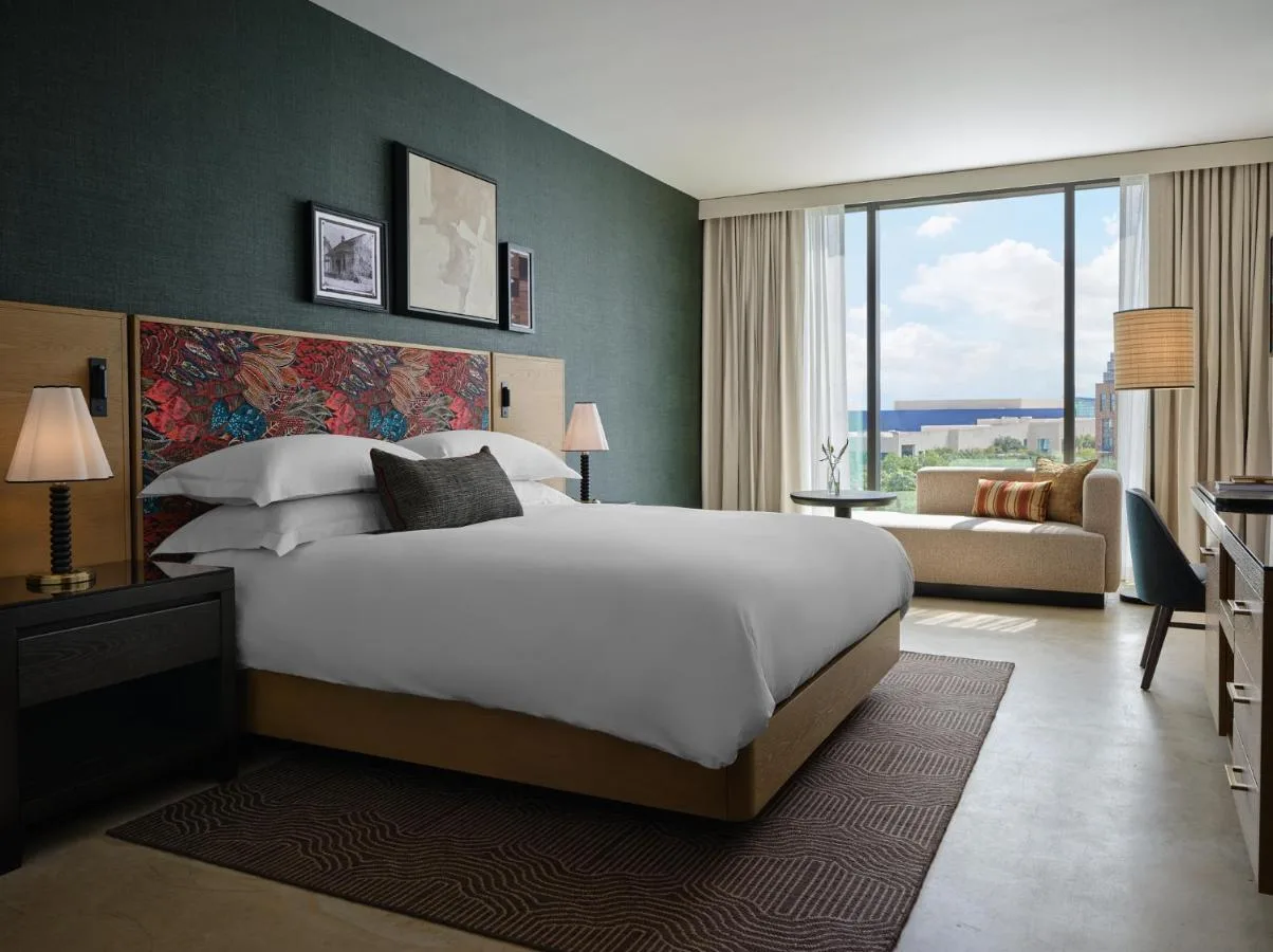 A modern hotel room featuring a king-size bed with a colorful patterned headboard, ambient lighting, a cozy seating area, and a large window with city views.