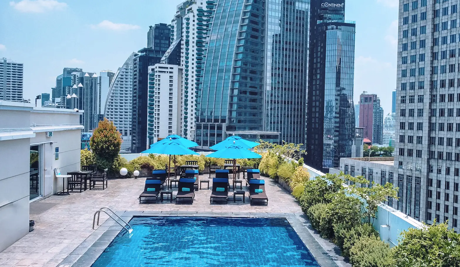 A New Bangkok Hotel With Rooftop Views And A Pool—Take A Look 10 Rooftop pool at NH Bangkok Asoke with sun loungers, blue umbrellas, and a backdrop of Bangkok’s modern skyscrapers.
