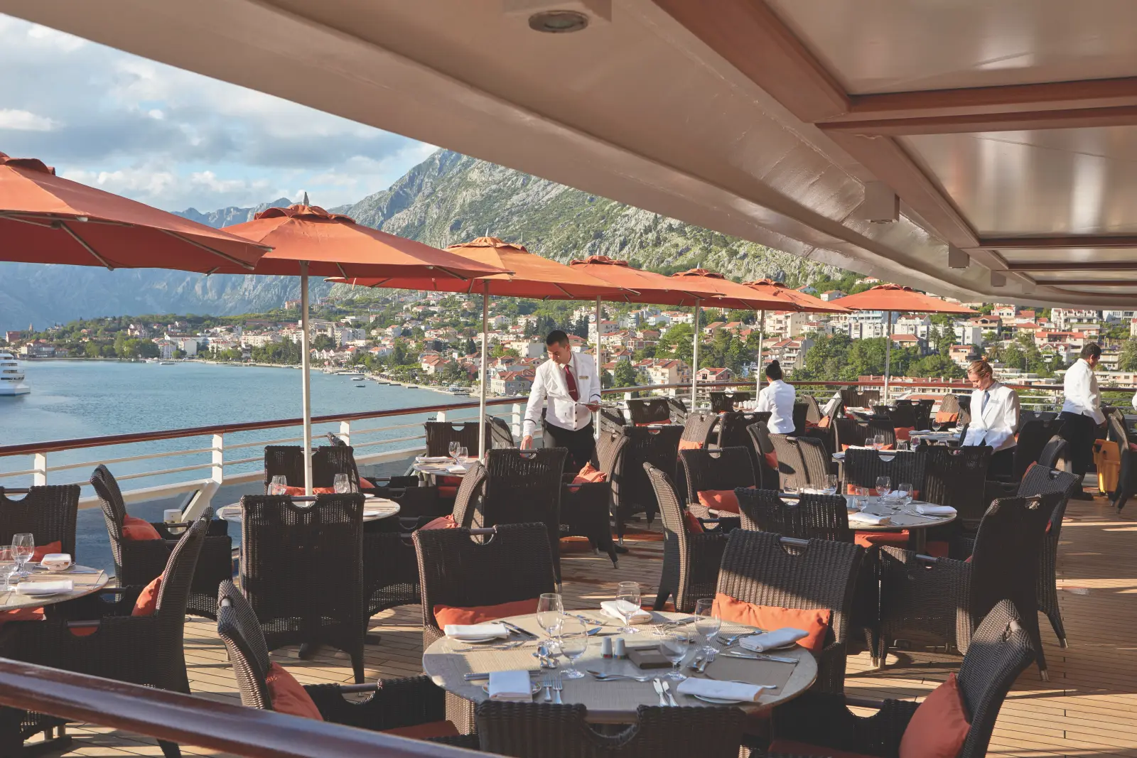 Outdoor dining area on an Oceania Cruises ship with stunning coastal and mountain views, featuring elegantly set tables and attentive service.