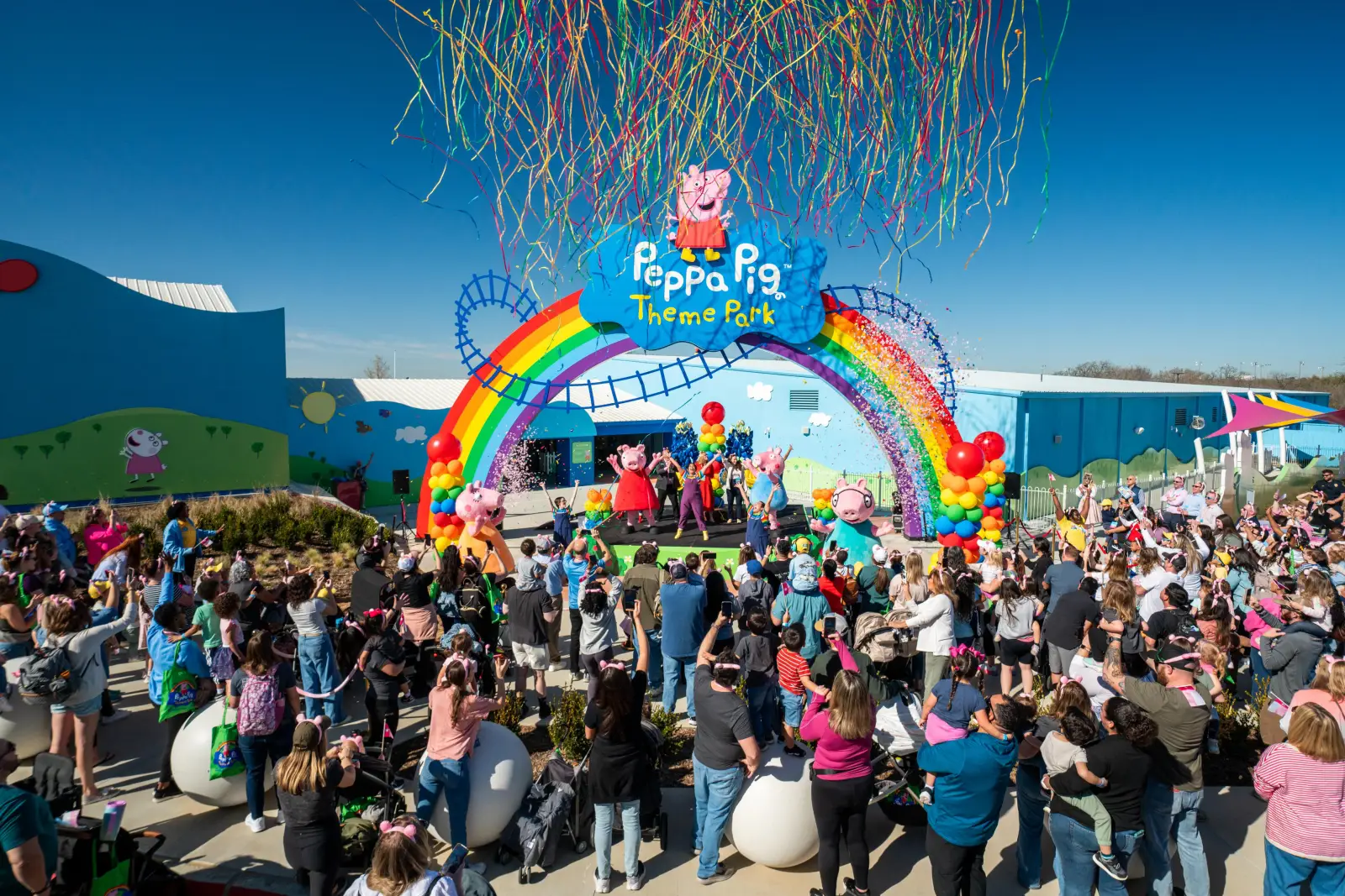 Grand opening celebration at Peppa Pig Theme Park Dallas-Fort Worth with a crowd cheering under a rainbow entrance as Peppa Pig and friends perform on stage.