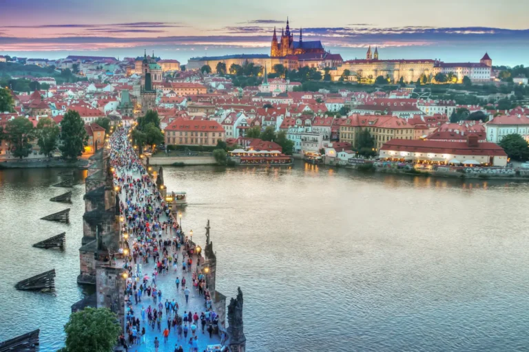 A stunning sunset view of Prague’s Charles Bridge filled with tourists, leading to Prague Castle, illuminated against the evening sky.