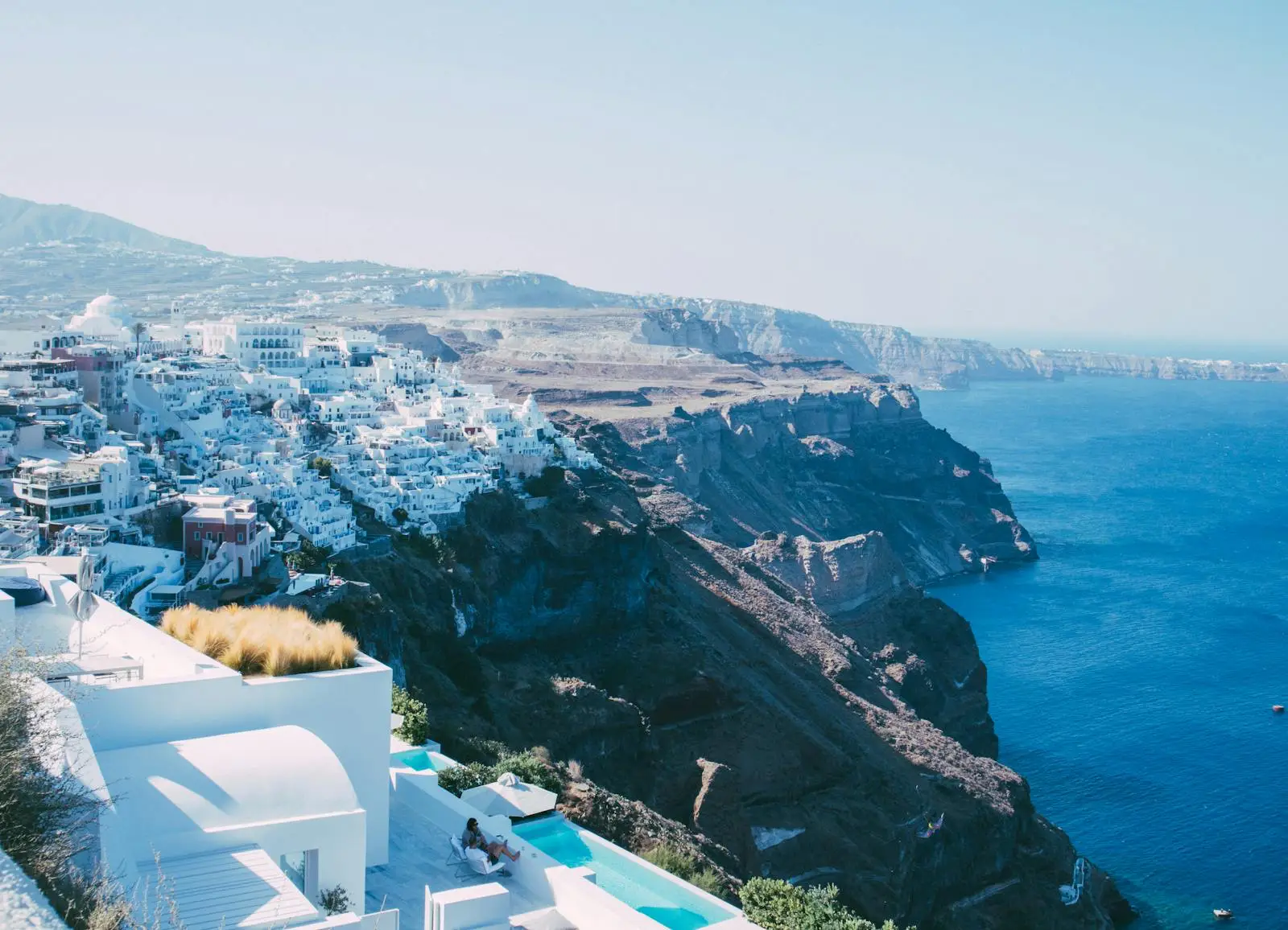 A view of Santorini’s white-washed buildings perched on cliffs overlooking the deep blue Aegean Sea, with rugged landscapes in the background.