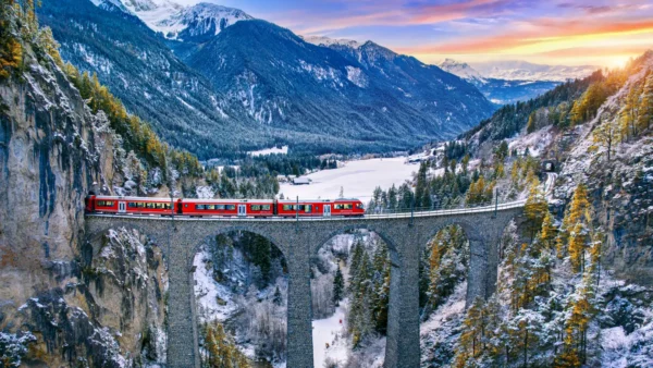 A red Swiss train crosses a historic stone viaduct in a scenic alpine landscape, surrounded by snow-covered mountains and evergreen forests at sunset.