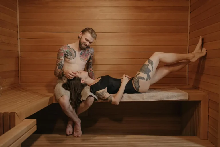 A tattooed couple relaxing in a wooden sauna, with the woman lying down and the man gently touching her head.