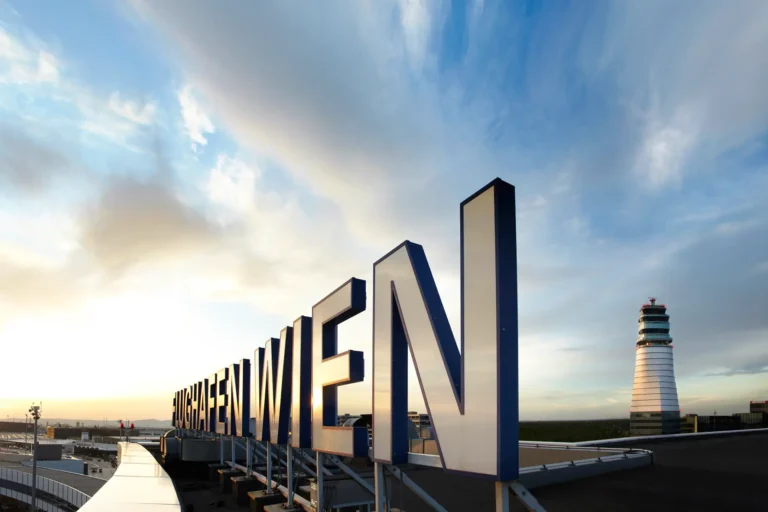 Large illuminated “Flughafen Wien” sign at Vienna Airport with the control tower in the background during sunset.