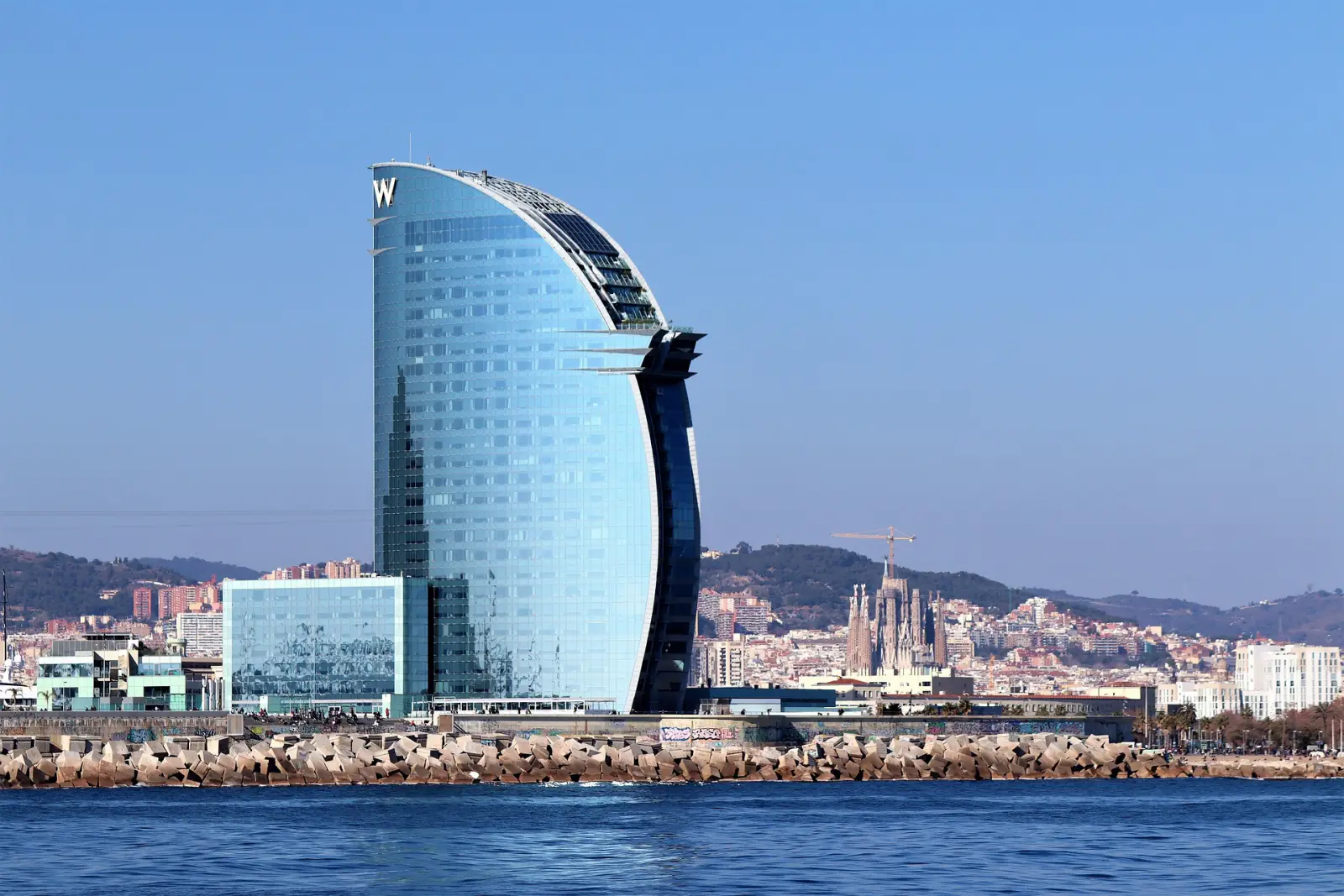 Best Places To Travel In Europe In August (2025 Edition) 3 The iconic W Barcelona hotel, a sail-shaped glass skyscraper on the Barcelona coastline, with the Sagrada Família and city skyline in the background.