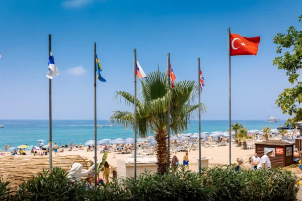 Flags of multiple countries, including Turkey, flying above Cleopatra Beach in Alanya, Turkey, with sunbathers and umbrellas along the coastline.