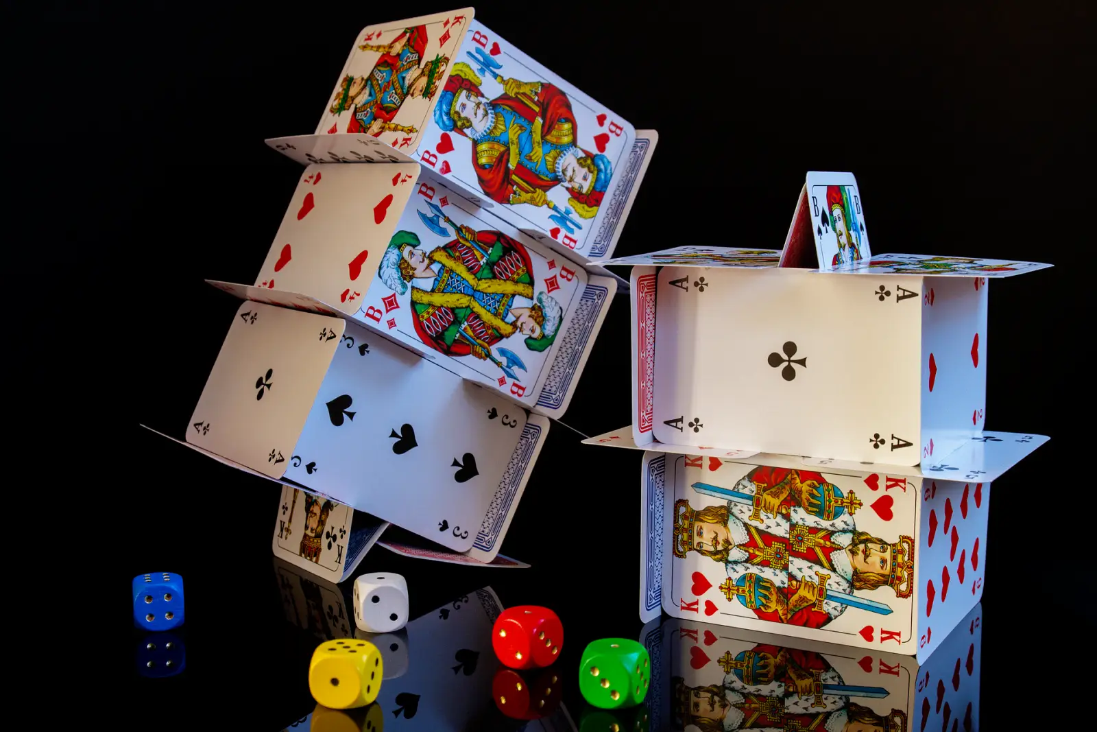 Two towers made of playing cards with colorful dice scattered on a reflective black surface