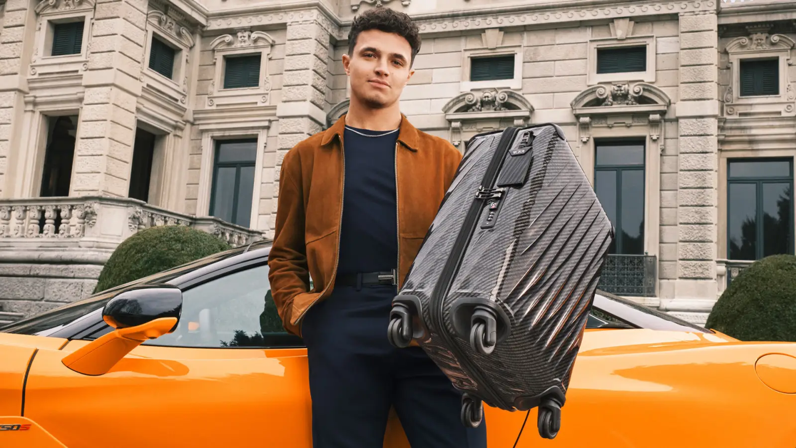 Lando Norris holding a TUMI 19 Degree Lite carry-on beside an orange sports car