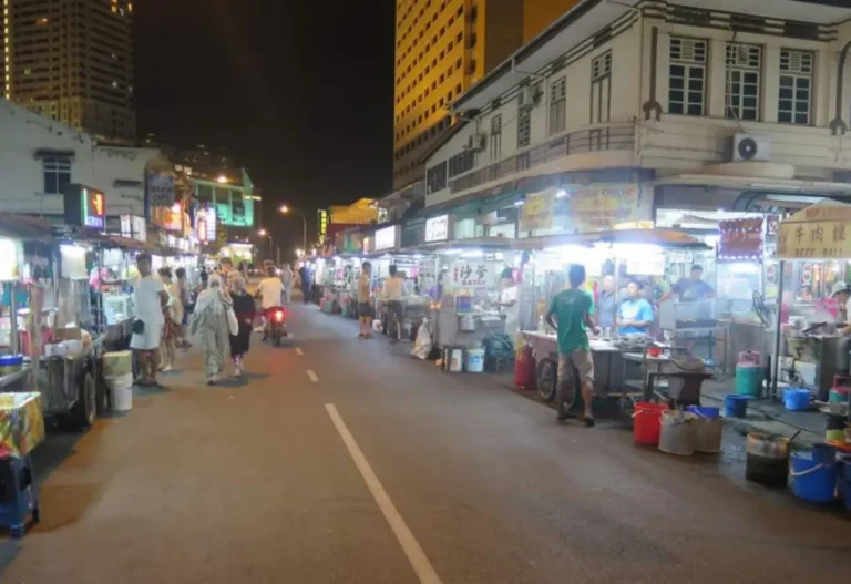 Night view of a busy Penang street food market with stalls, vendors, and visitors walking along the road