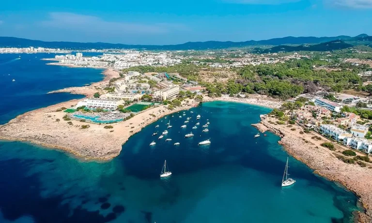 Aerial view of Port des Torrent beach, a cove with turquoise waters and anchored boats on the west coast of Ibiza, Spain.