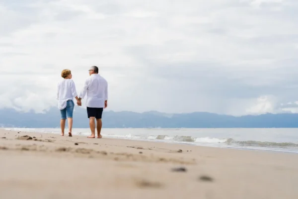 Senior couple walking hand in hand along a peaceful beach with mountains in the distance.