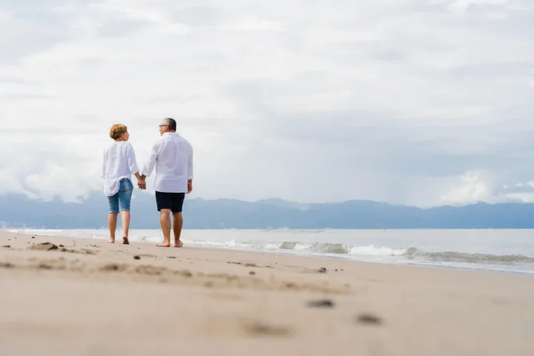Senior couple walking hand in hand along a peaceful beach with mountains in the distance.