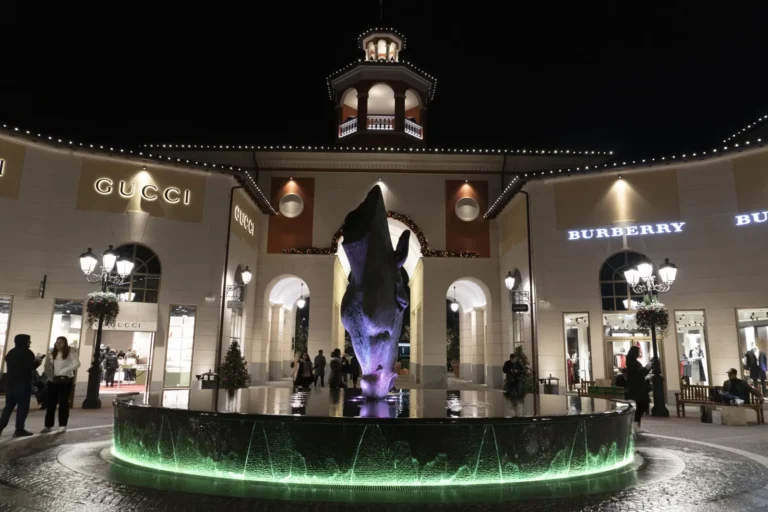 Night view of Gucci and Burberry storefronts at Serravalle Designer Outlet with illuminated fountain and sculpture