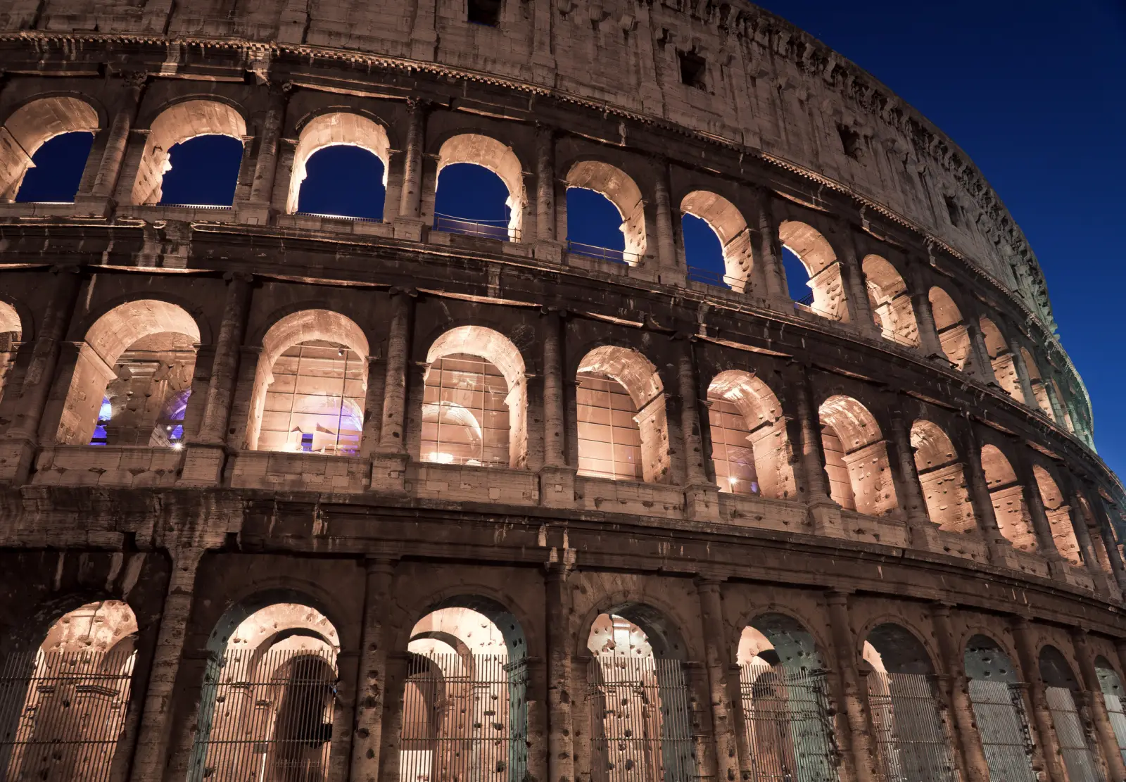 Why U.s. Travelers Are Flocking To Colosseum Night Tours In Rome 2 The Colosseum illuminated at night, showcasing its arches and stone facade against a deep blue sky.