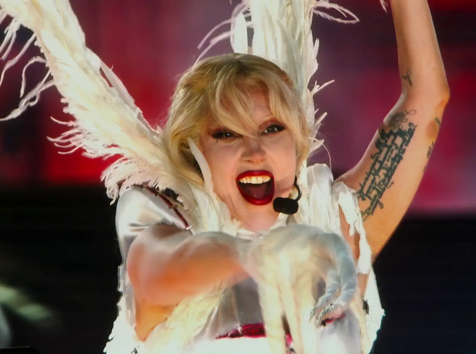 Lady Gaga performing on stage in a white feathered costume, singing energetically with a headset microphone.