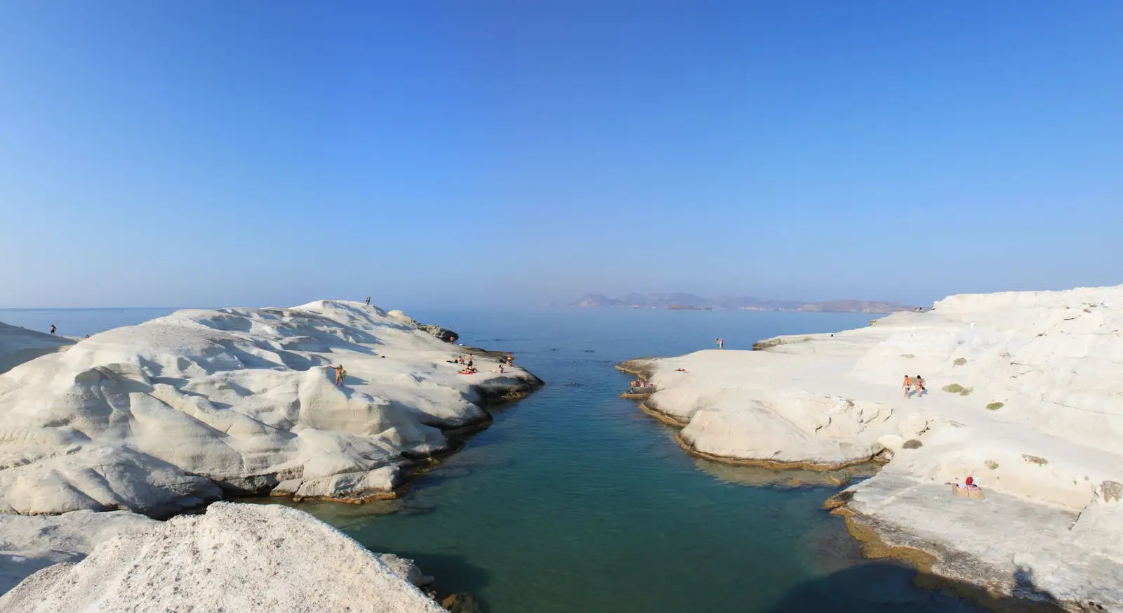 Santorini Battles Booking Drop — Here’s Where Travelers Are Going Instead 3 People relaxing on the white rock formations at Sarakiniko Beach in Milos, Greece, with clear blue sky and calm sea.