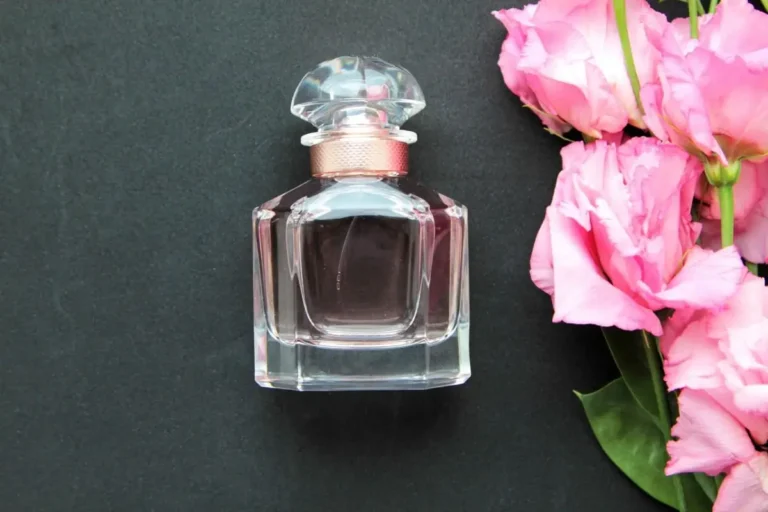 Glass perfume bottle with pink cap next to fresh pink flowers on a dark background