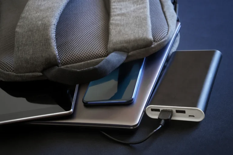 Technology 21 Power bank charging electronic devices partially hidden under a backpack with a phone, tablet, and laptop.