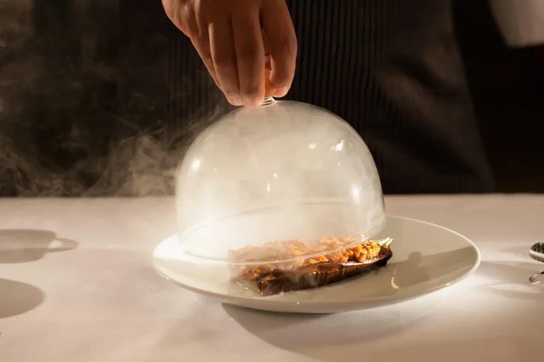 Chef lifting a glass cloche releasing aromatic smoke over a stuffed roasted eggplant on a white plate in a fine-dining setting