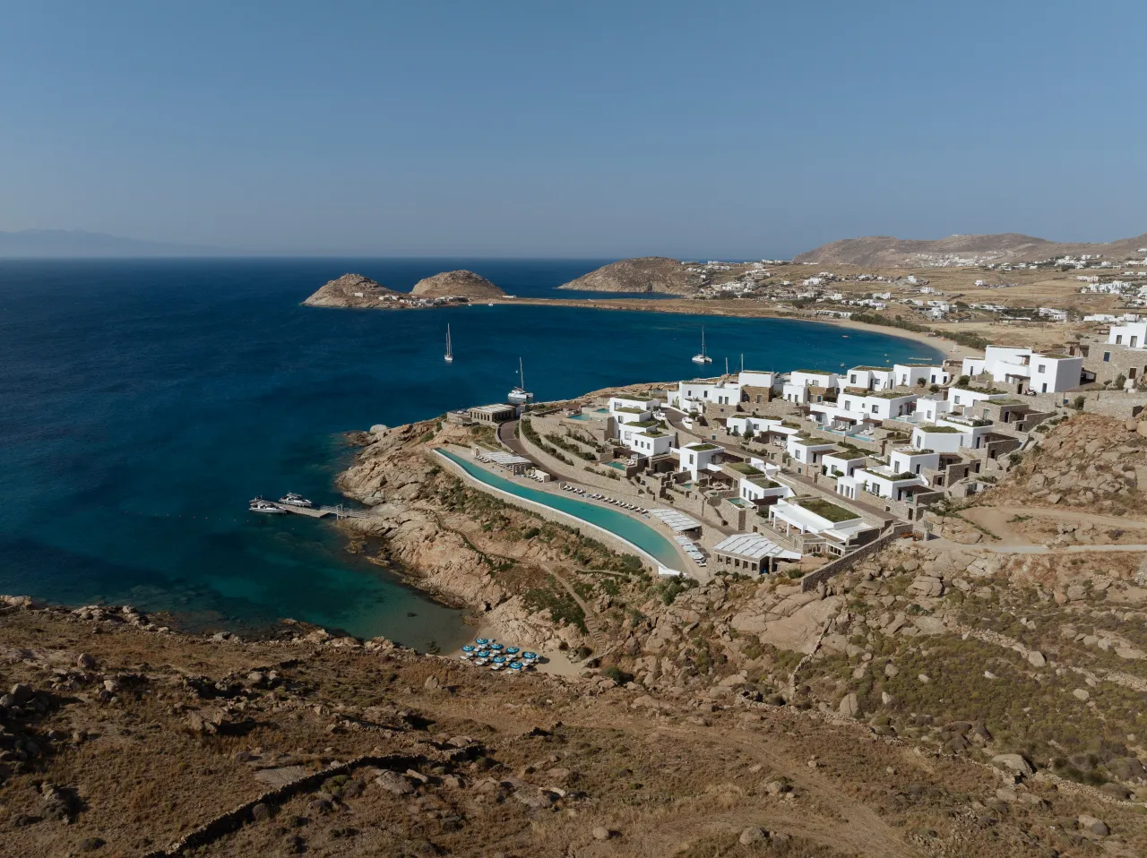 Aerial view of Cali Mykonos resort on Greece’s coastline, with whitewashed villas, an expansive infinity pool, rocky hills, and the blue Aegean Sea.