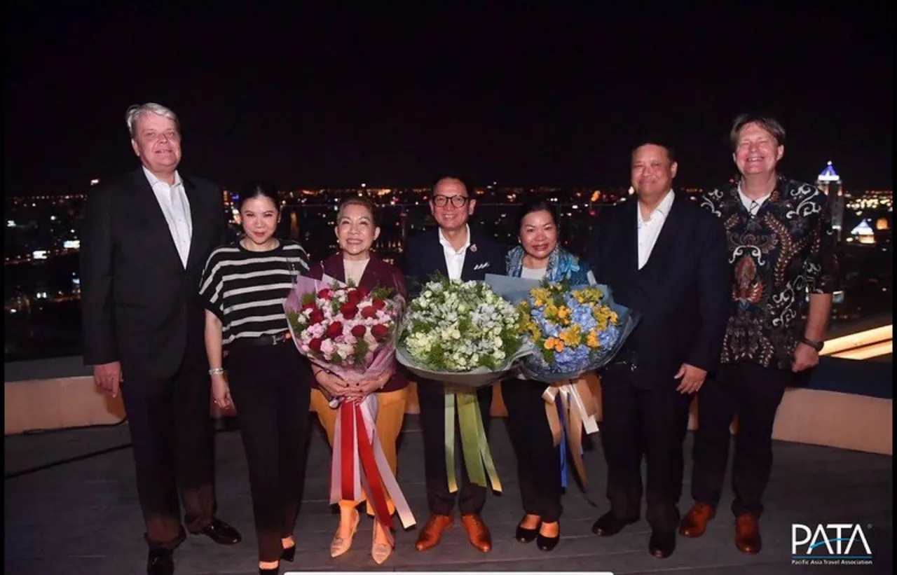 Group photo at night of PATA event attendees in Bangkok, with leaders holding flower bouquets and the city skyline in the background.