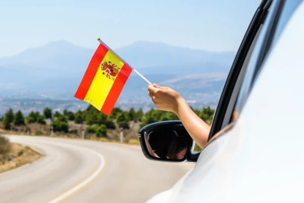 Person holding a Spanish flag out of a car window while driving along a winding road with mountains in the background.