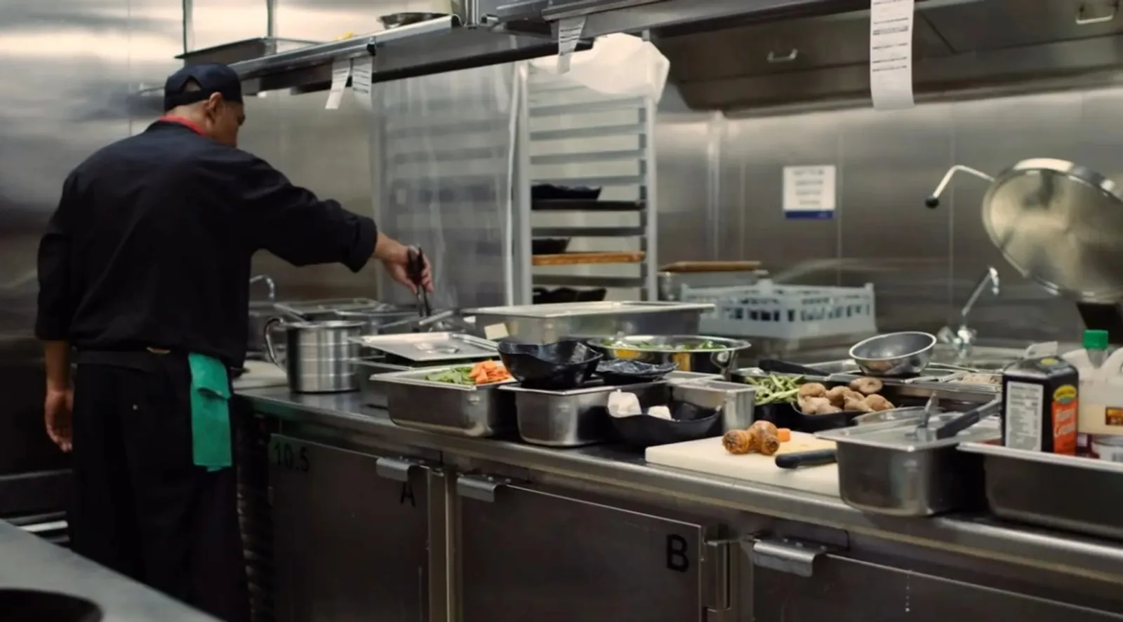 Behind-The-Scenes Cruise Tour: Holland America Unlocks Ship Secrets 4 Cruise ship chef preparing food in a stainless-steel galley with fresh ingredients and cooking utensils spread across the counter.