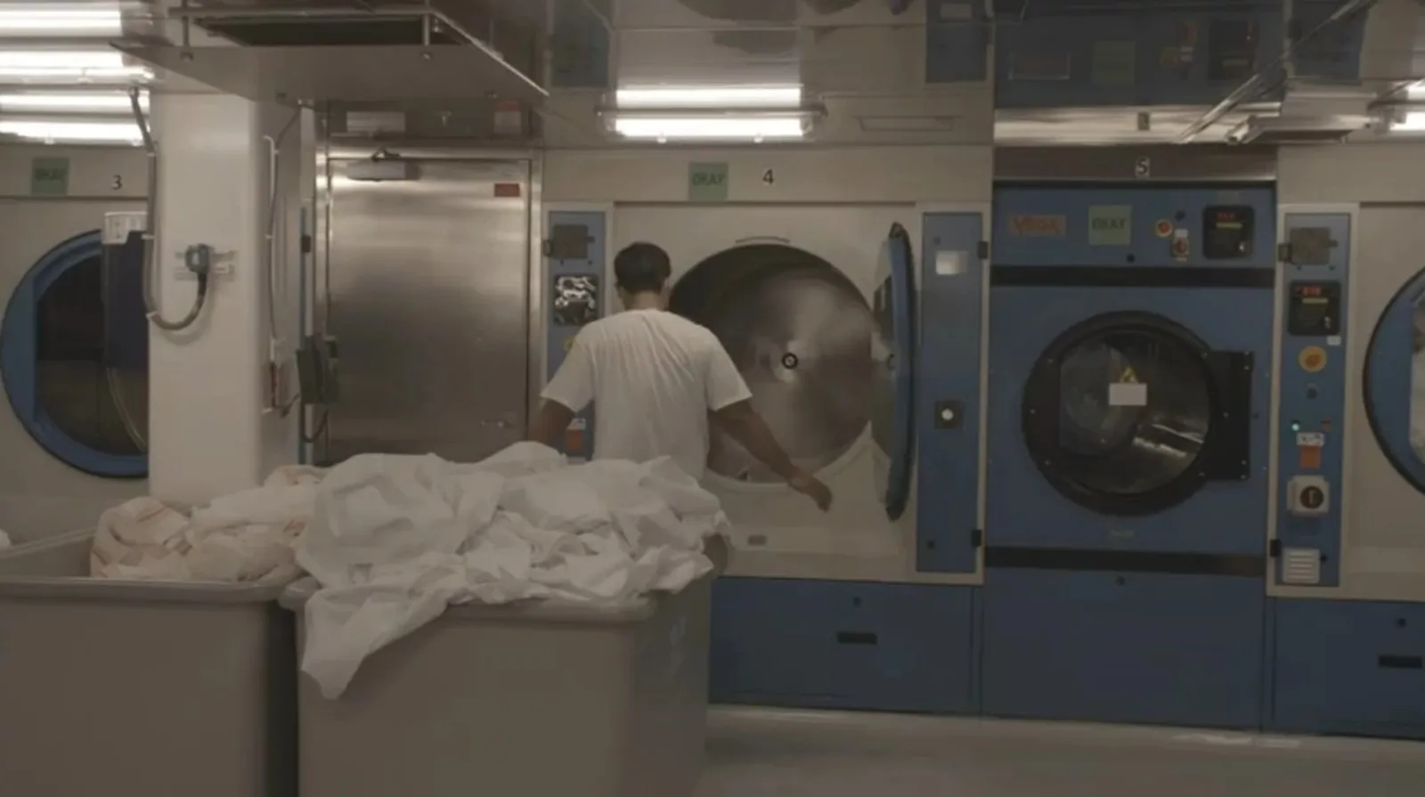 Behind-The-Scenes Cruise Tour: Holland America Unlocks Ship Secrets 5 Crew member operating an industrial washing machine in the cruise ship’s laundry room, with large bins of white linens in front.