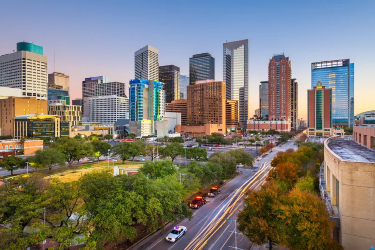 Downtown Houston skyline at sunset with tall modern buildings, green trees, and light trails from moving traffic.