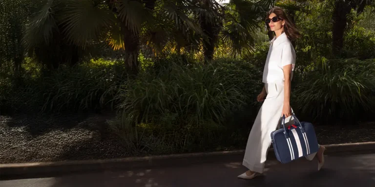 Woman walking confidently along a palm-lined path, wearing a white outfit and carrying a navy La Croisette laptop bag by Air France with white stripes.