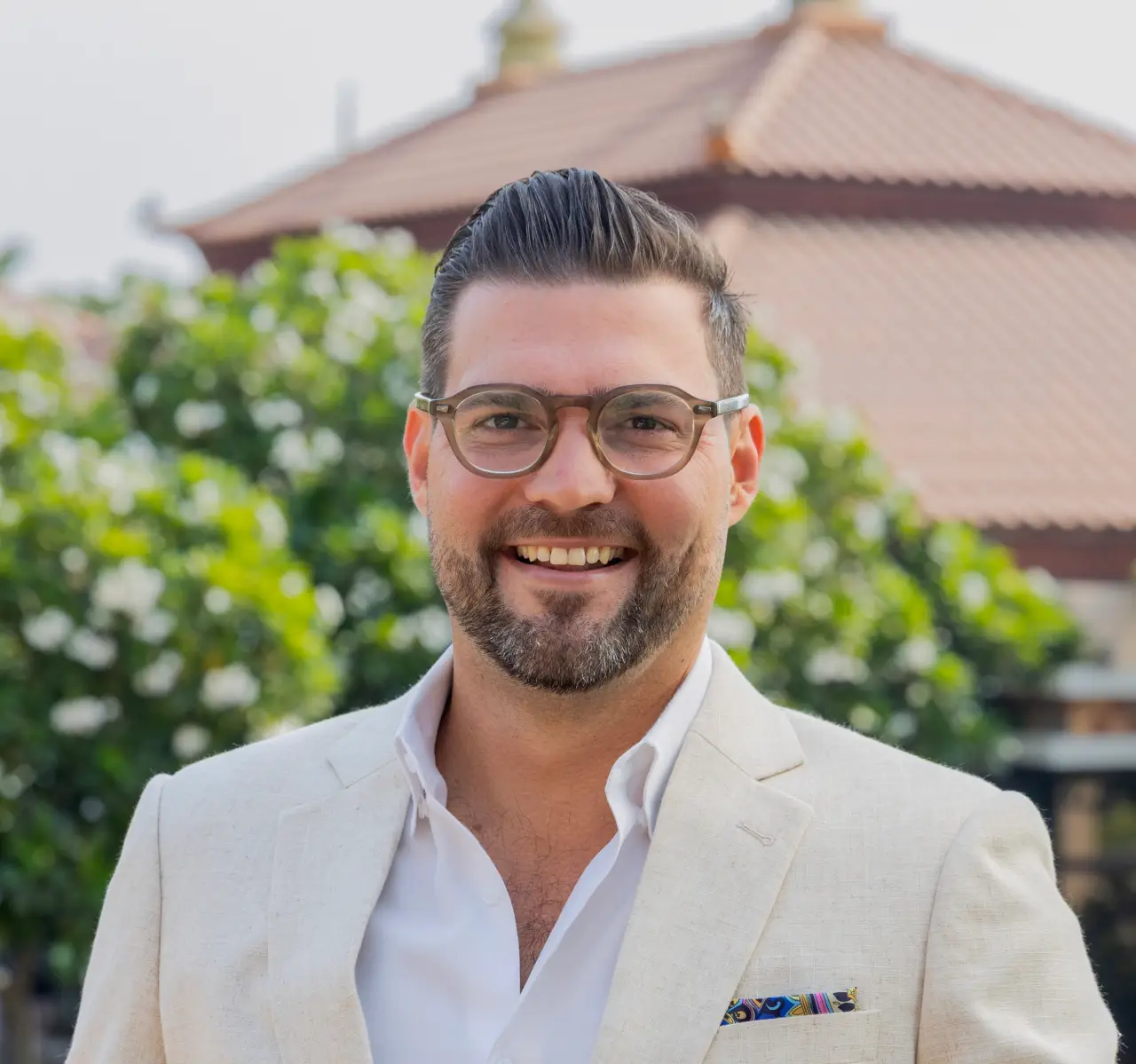 Micheal Robinson in a light beige suit with glasses, standing outdoors in front of greenery and tiled-roof buildings.