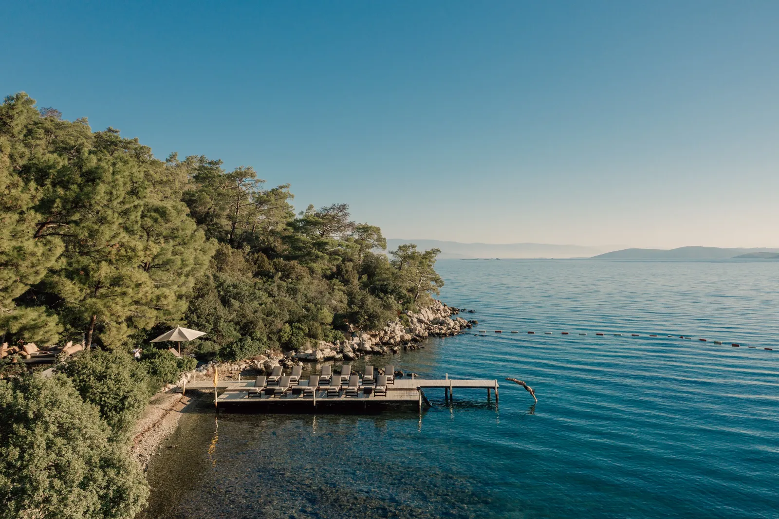 Secluded Bodrum coastline with a wooden pier, sun loungers, lush greenery and clear Aegean waters at the planned Aman Residences site.