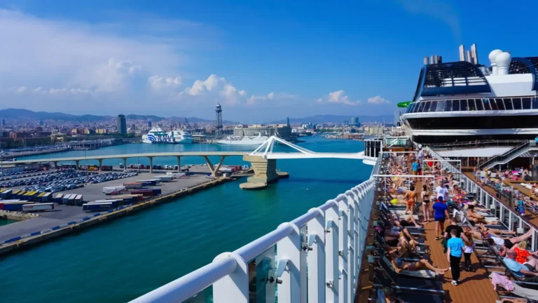 Cruise ship passengers sunbathing on deck as the vessel docks in the Port of Barcelona, with city skyline and harbor infrastructure visible in the background.