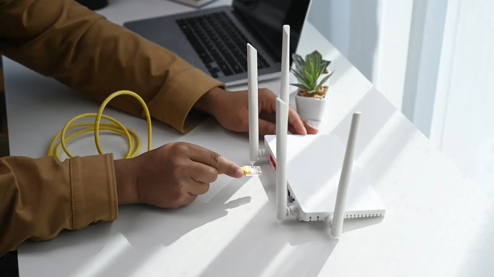 Top Tips For Choosing The Right Internet Plan 2 Person plugging an ethernet cable into a wireless router next to a laptop on a white desk.